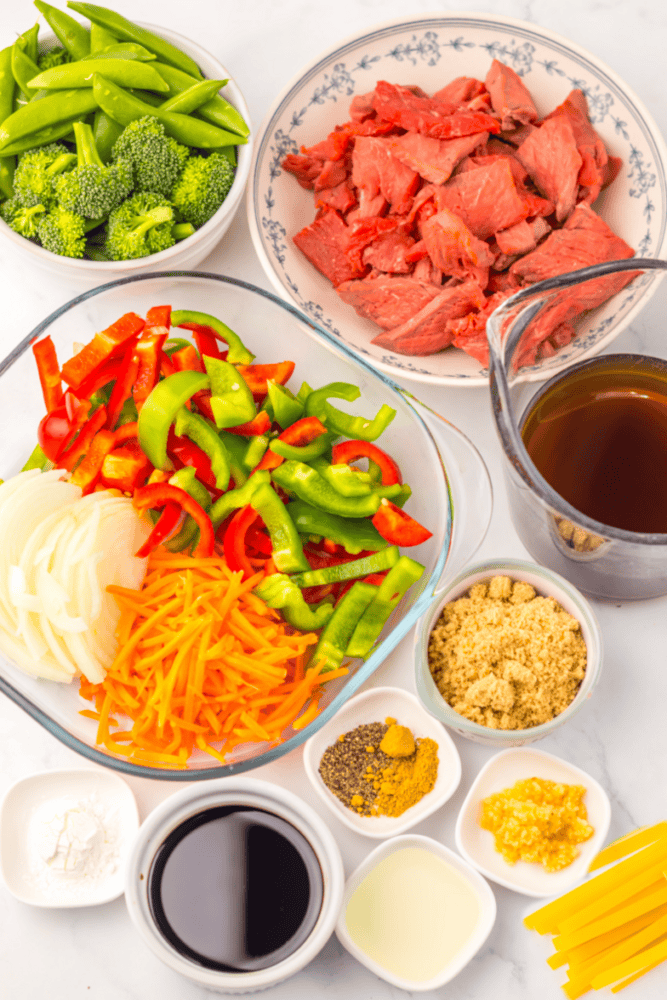 Bowls of sliced beef, mixed vegetables, broth, seasonings, and noodles arranged on a white surface for stir-fry.