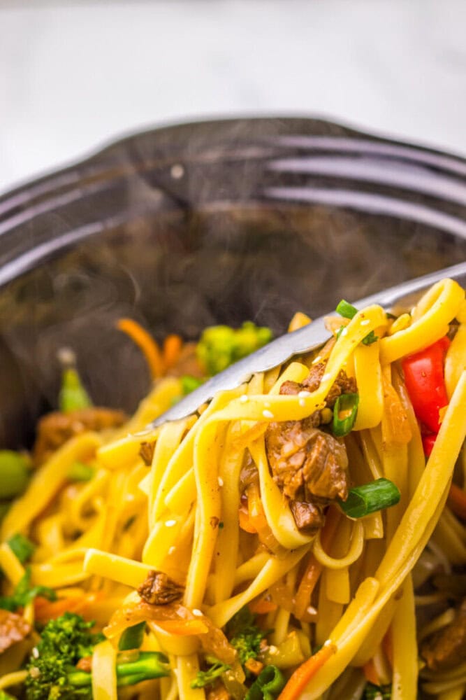 Close-up of tongs lifting noodles with beef, vegetables, and sesame seeds from a black bowl.