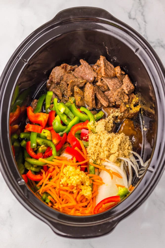 Sliced beef, bell peppers, carrots, onions, garlic, and brown sugar in a slow cooker before mixing.