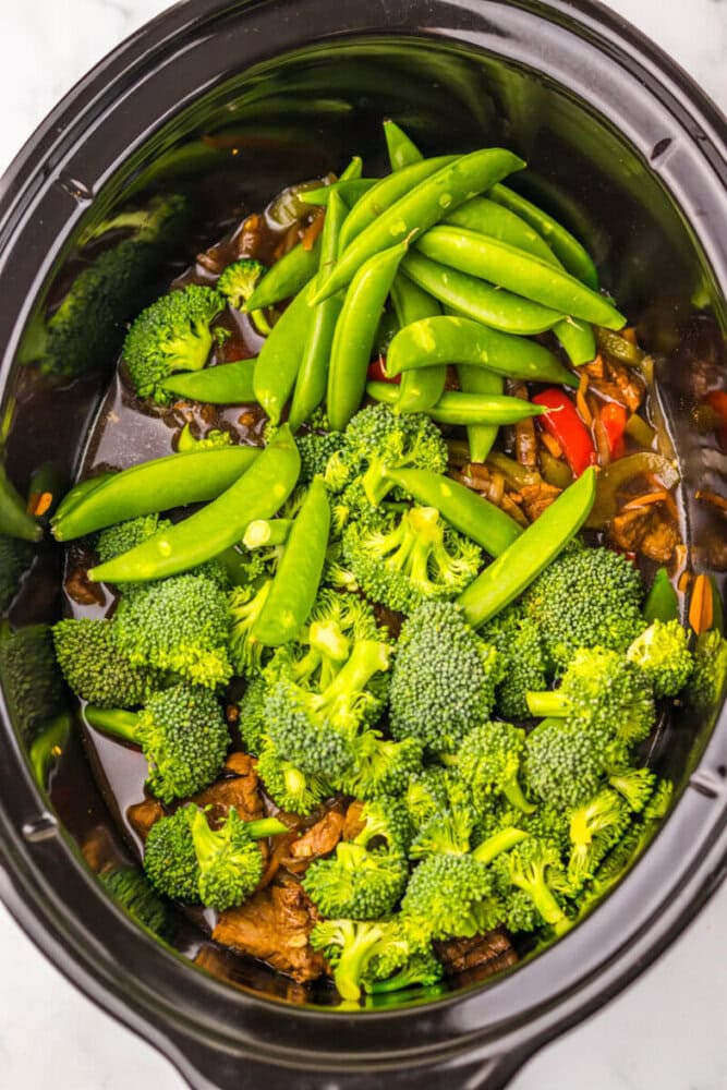 Broccoli and snap peas on top of meat and vegetables in a slow cooker, ready to be cooked.