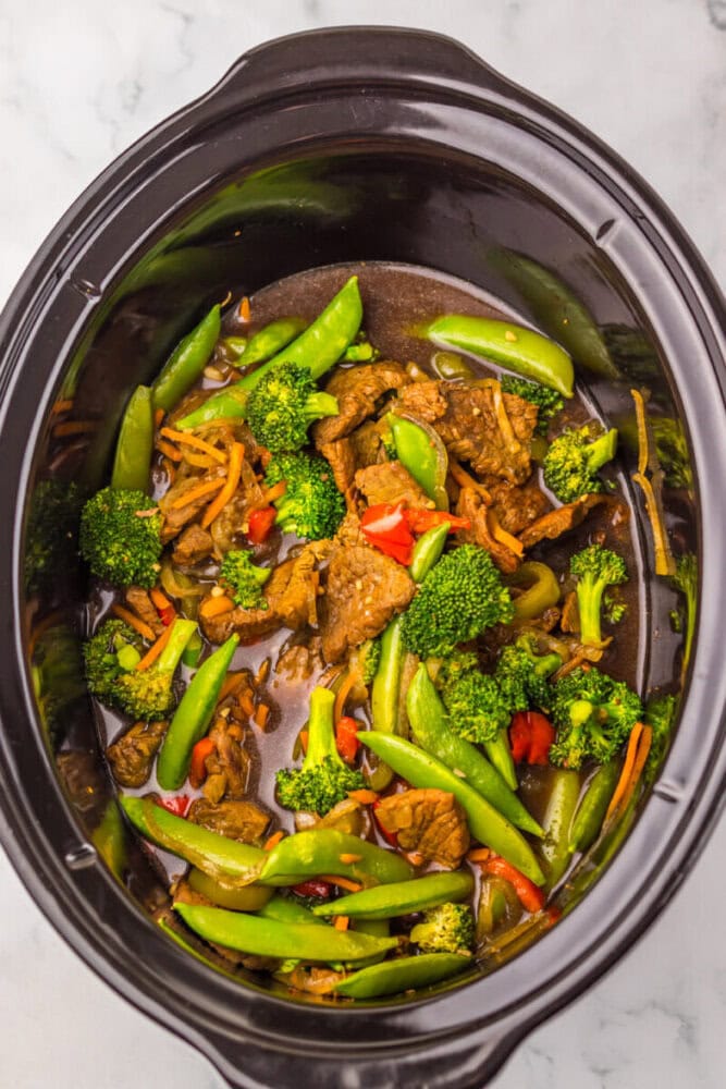 Beef and mixed vegetables in a dark slow cooker with broccoli, snap peas, carrots, and red bell peppers.