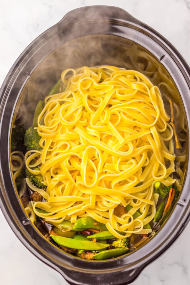 A slow cooker filled with uncooked fettuccine noodles and mixed vegetables, steaming hot.
