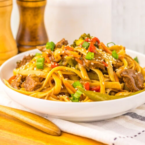 A bowl of beef stir-fry noodles with vegetables, topped with sesame seeds and green onions.