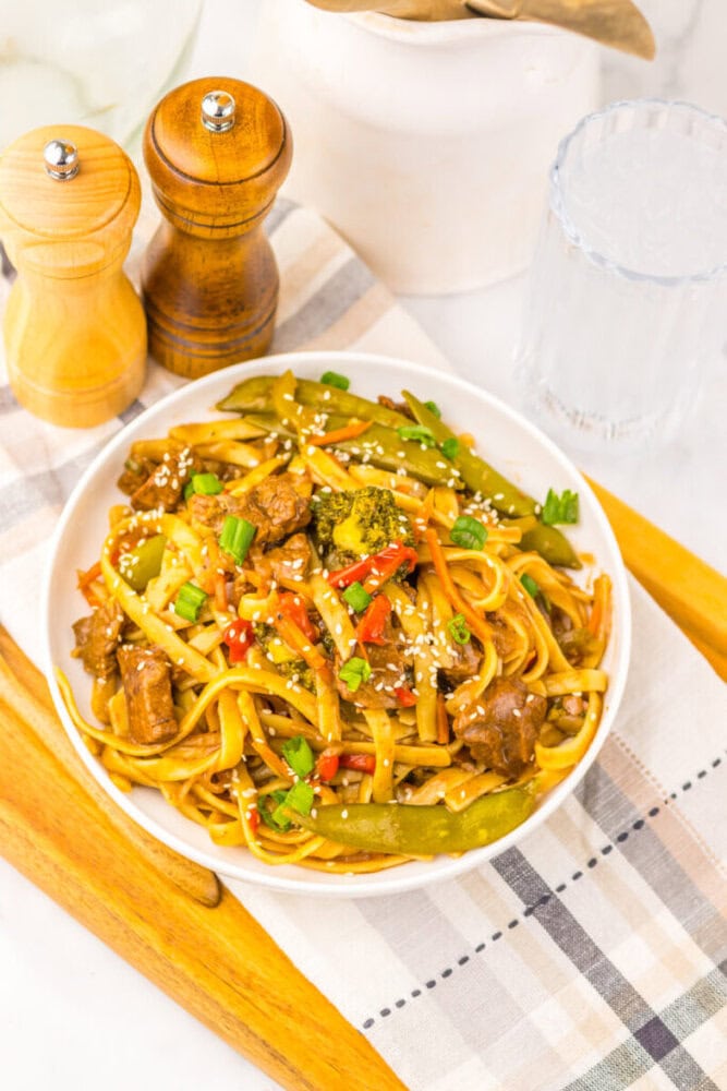 A bowl of stir-fried noodles with beef, vegetables, sesame seeds, and green onions on a plaid napkin.