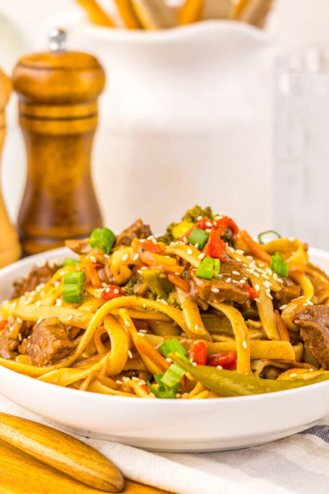 A bowl of beef lo mein noodles with vegetables, topped with sesame seeds and chopped green onions.