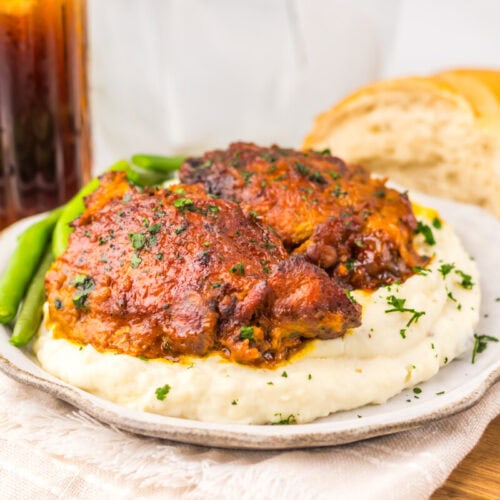 Two pieces of glazed chicken on mashed potatoes with green beans, bread, and a glass of soda in the background.