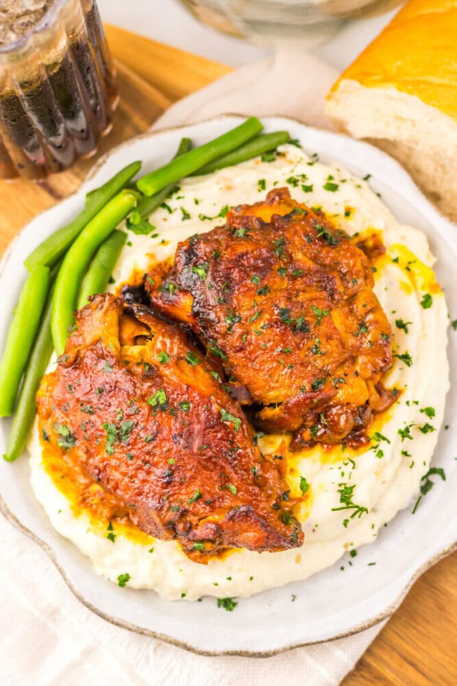 Two roasted chicken thighs on mashed potatoes, garnished with parsley, served with green beans and a drink.