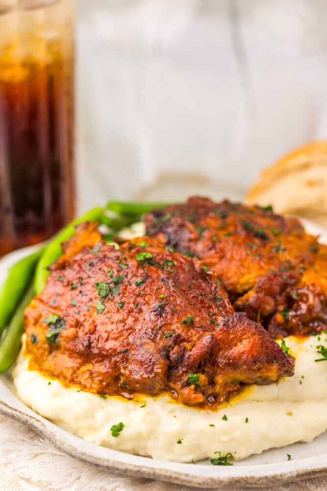 Barbecue chicken thighs on mashed potatoes, garnished with herbs, served with green beans and a drink in the background.