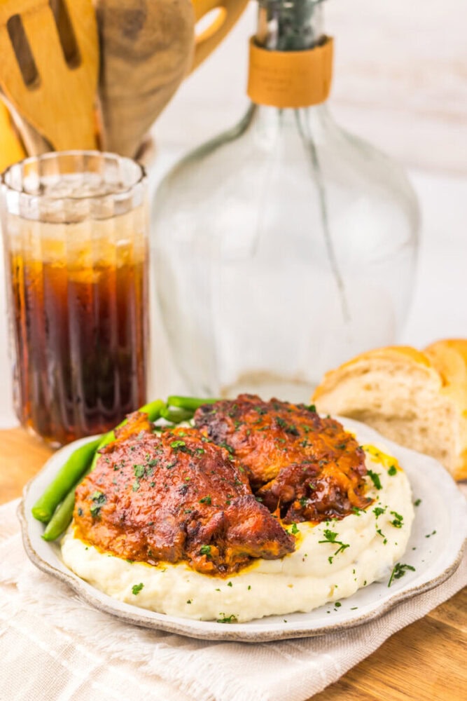 Plate of BBQ chicken on mashed potatoes with green beans, a glass of soda, and bread in the background.