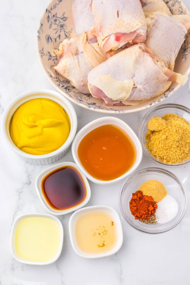 Raw chicken pieces in a bowl with small bowls of mustard, honey, brown sugar, spices, and sauces on a white surface.