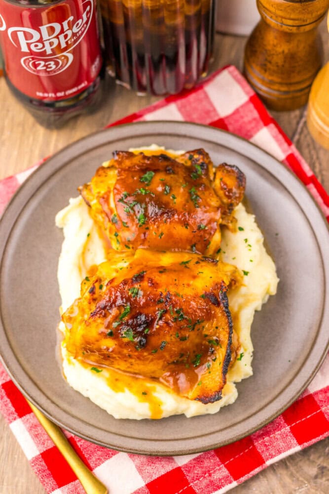 Two glazed chicken thighs on mashed potatoes, served on a plate with a red checkered napkin and drink nearby.