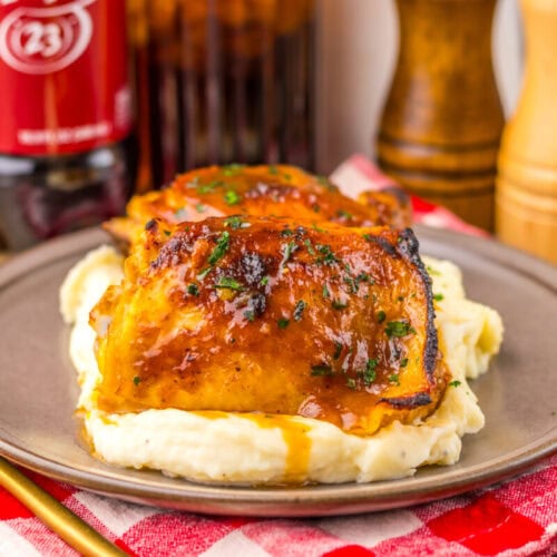 Baked chicken thighs on mashed potatoes, served on a plate with sauce and herbs, on a red plaid cloth.