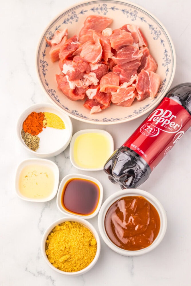 Raw pork chunks, Dr Pepper bottle, and various spices and sauces in bowls arranged on a white surface.