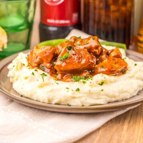 Mashed potatoes topped with beef stew on a plate, with bread, a drink, and condiments in the background.