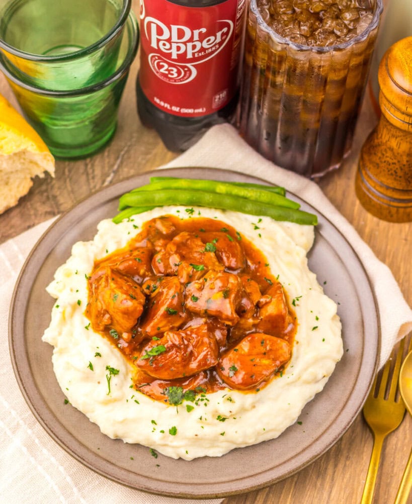 Plate of mashed potatoes topped with brown gravy and meat, with green beans, bread, and a Dr Pepper drink.