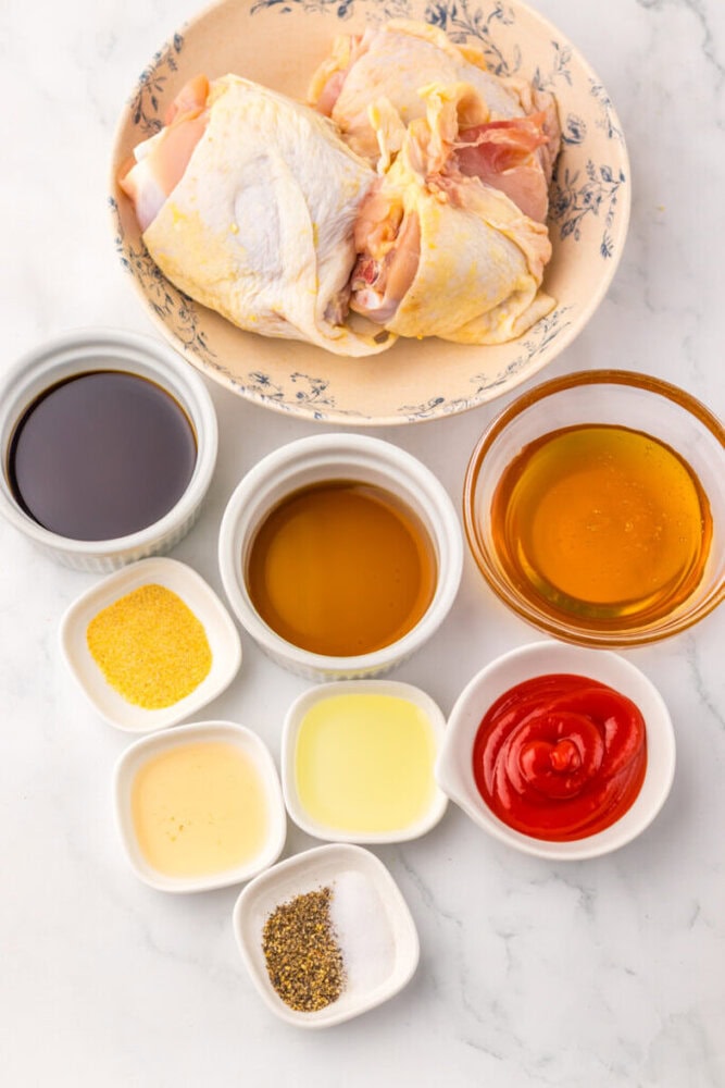 Raw chicken, sauces, seasonings, and spices arranged in bowls on a white surface, viewed from above.