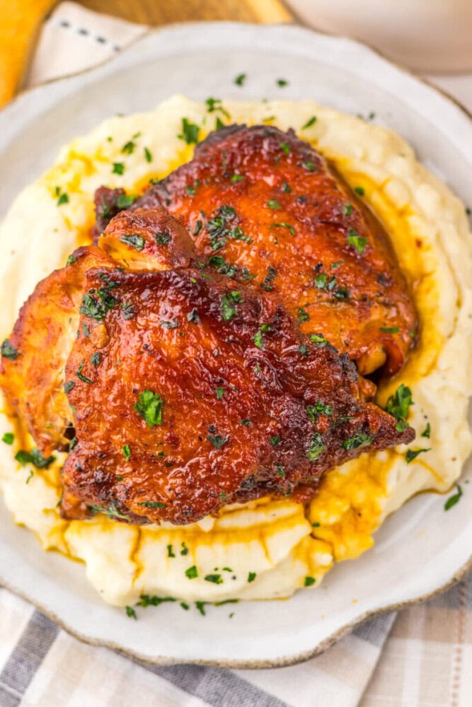Two pieces of glazed roasted chicken served on a bed of creamy mashed potatoes, garnished with chopped herbs.