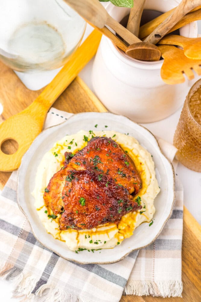 Plate of glazed roasted chicken thighs on mashed potatoes, garnished with herbs, on a plaid napkin with utensils nearby.