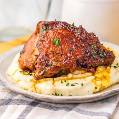 Barbecue chicken thigh on mashed potatoes, garnished with herbs, served on a plate over a checkered cloth.