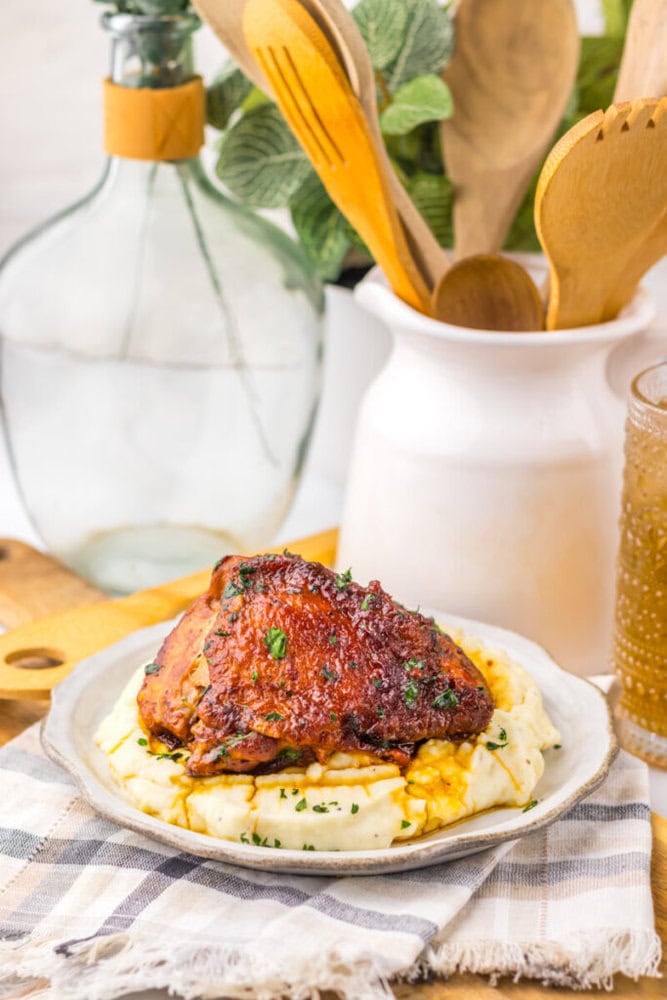 A glazed chicken thigh on mashed potatoes, garnished with herbs, on a plate beside a drink and wooden utensils.