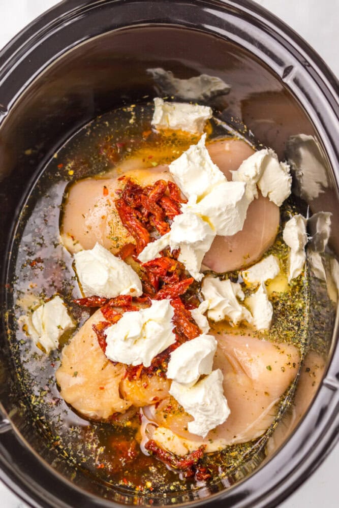 Raw chicken breasts, sun-dried tomatoes, and cream cheese in a slow cooker with herbs and seasonings.
