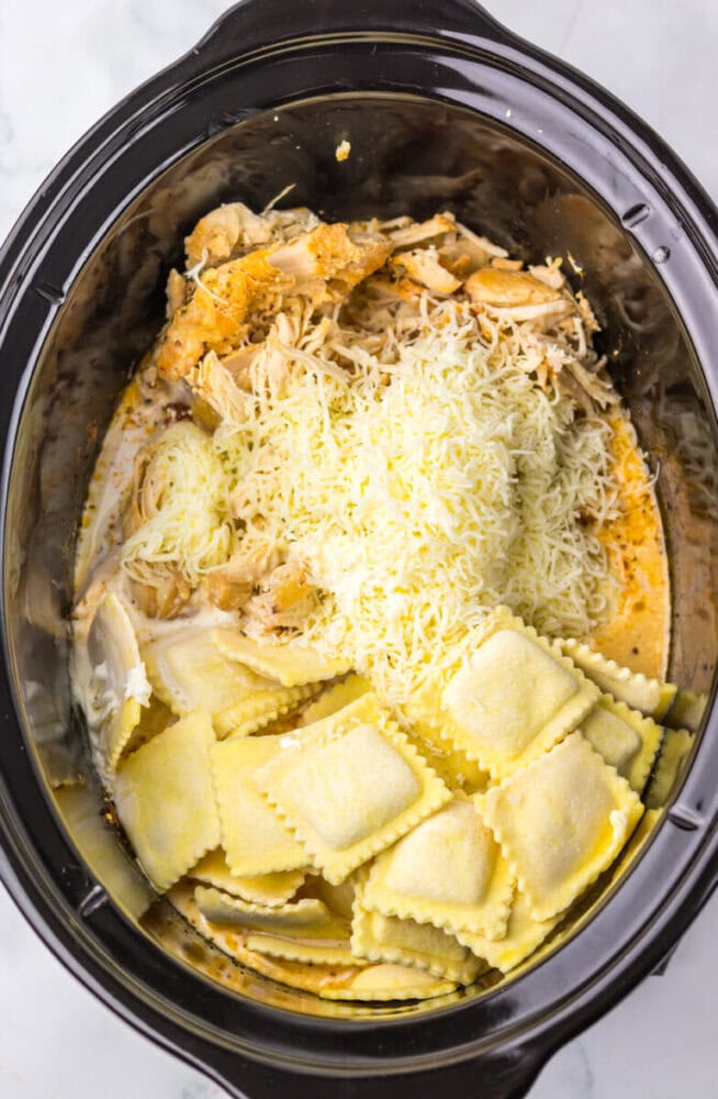 Shredded chicken, shredded cheese, and uncooked ravioli inside a black slow cooker.