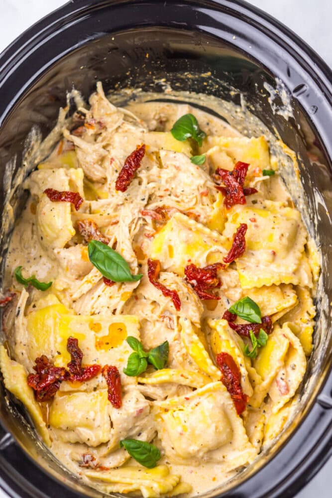 Creamy ravioli pasta with shredded chicken, sun-dried tomatoes, and basil leaves in a black slow cooker.
