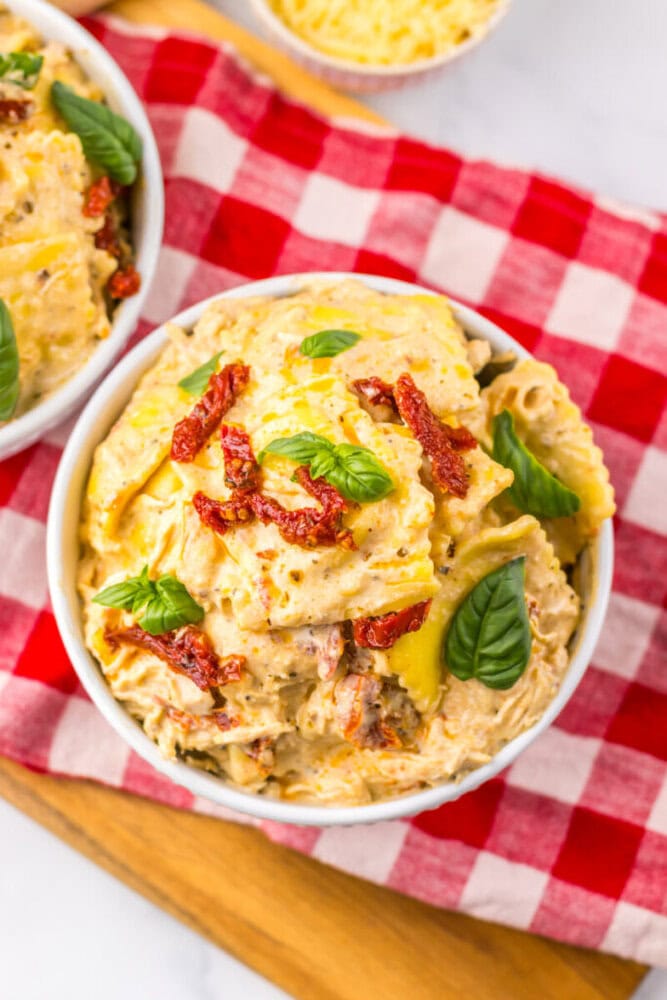 Bowl of creamy pasta with sun-dried tomatoes and basil on a red checkered cloth.