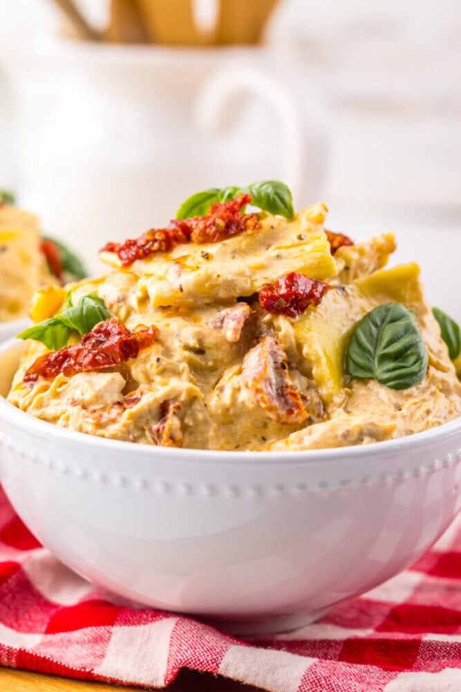 A white bowl of creamy pasta salad with sun-dried tomatoes, artichokes, and fresh basil on top.