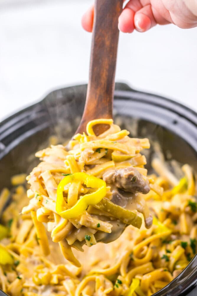 A wooden spoon lifts creamy beef and noodle pasta with yellow peppers from a black pot.