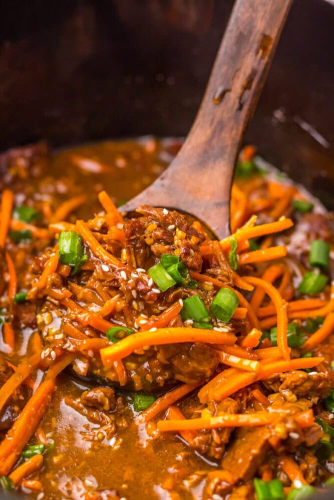 Close-up of a wooden spoon lifting beef stir fry with carrots, green onions, and sesame seeds in sauce.