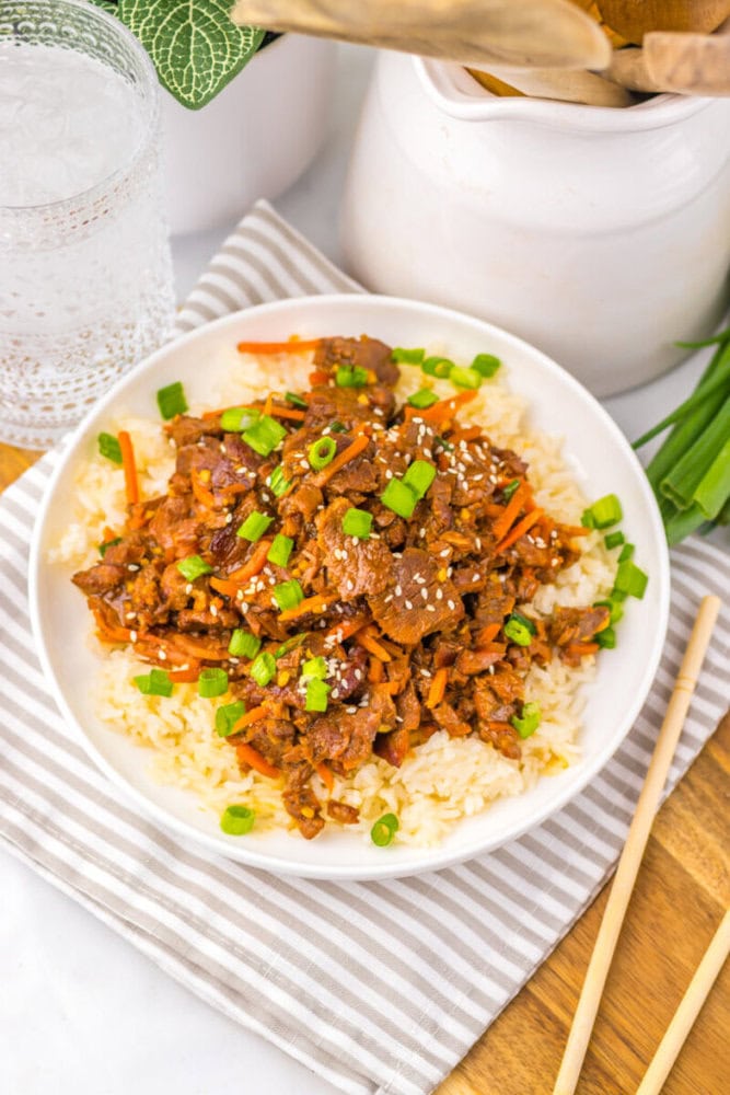 A bowl of rice topped with beef stir-fry, sesame seeds, and chopped green onions, next to chopsticks.