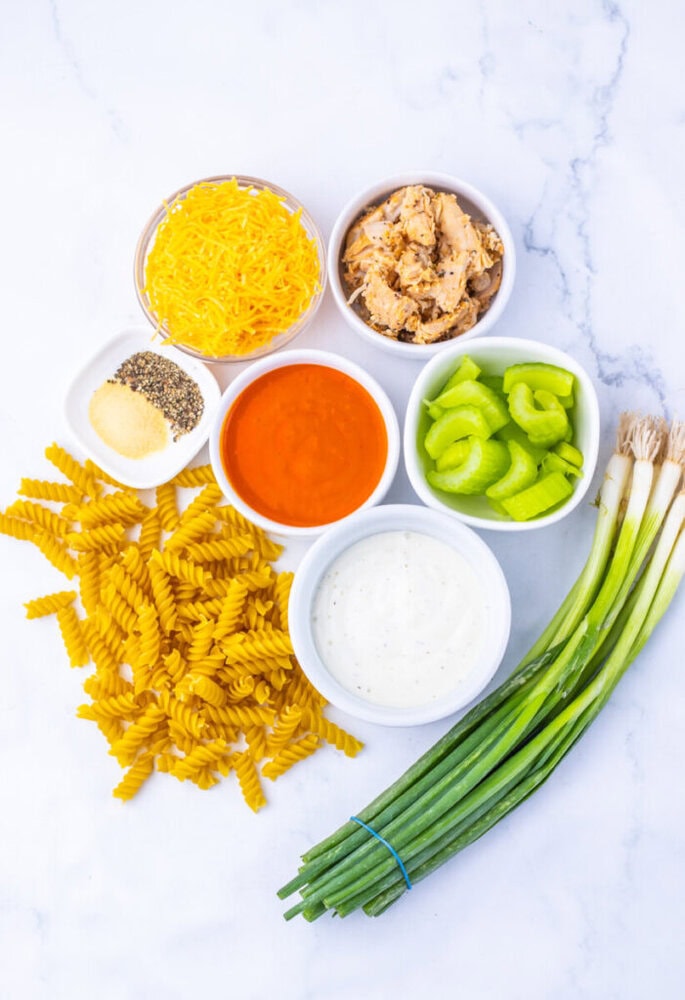 Dry pasta, cheese, shredded chicken, celery, green onions, ranch, buffalo sauce, and seasonings on a marble surface.