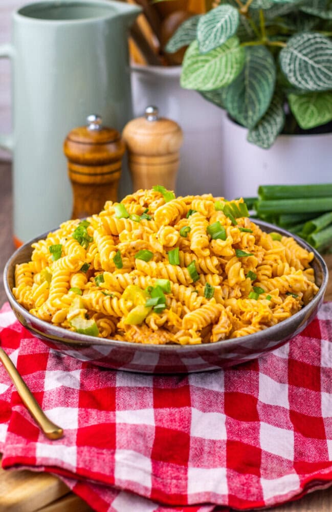 A bowl of creamy rotini pasta with green onions on a red checkered cloth, surrounded by kitchen items.