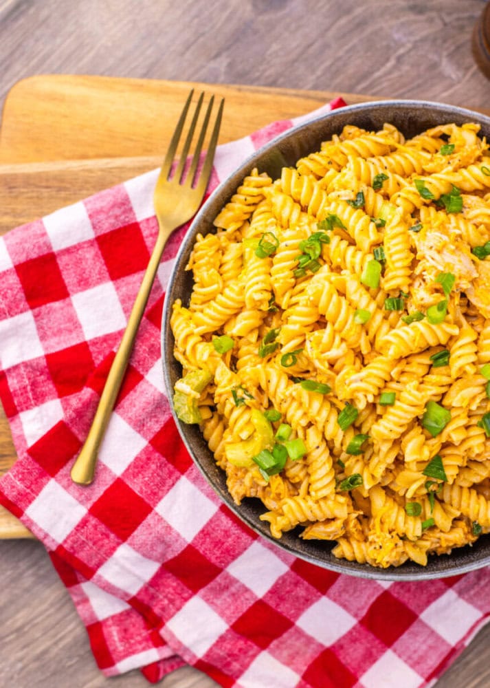 A bowl of creamy rotini pasta topped with chopped green onions on a red checkered napkin with a gold fork.