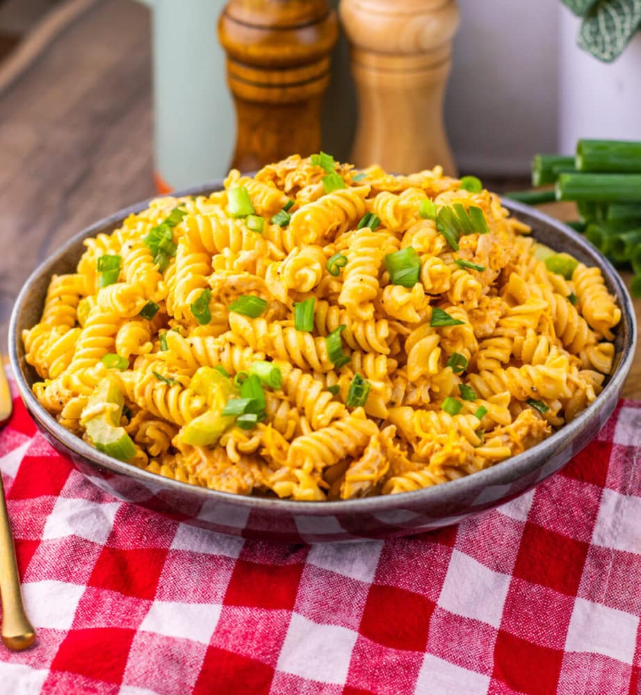 A bowl of cheesy rotini pasta garnished with chopped green onions on a red and white checkered cloth.