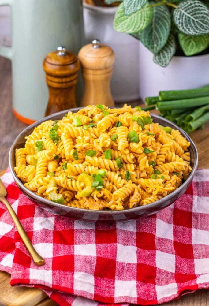 A bowl of rotini pasta with cheese and green onions on a red and white checkered cloth, with a gold fork nearby.