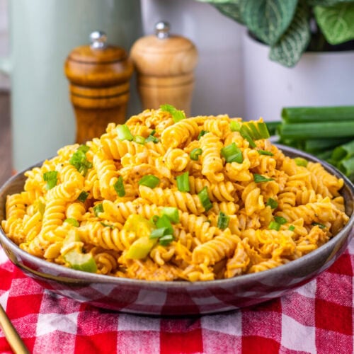 A bowl of creamy rotini pasta with green onions on a red plaid napkin, fork nearby, and salt and pepper shakers.
