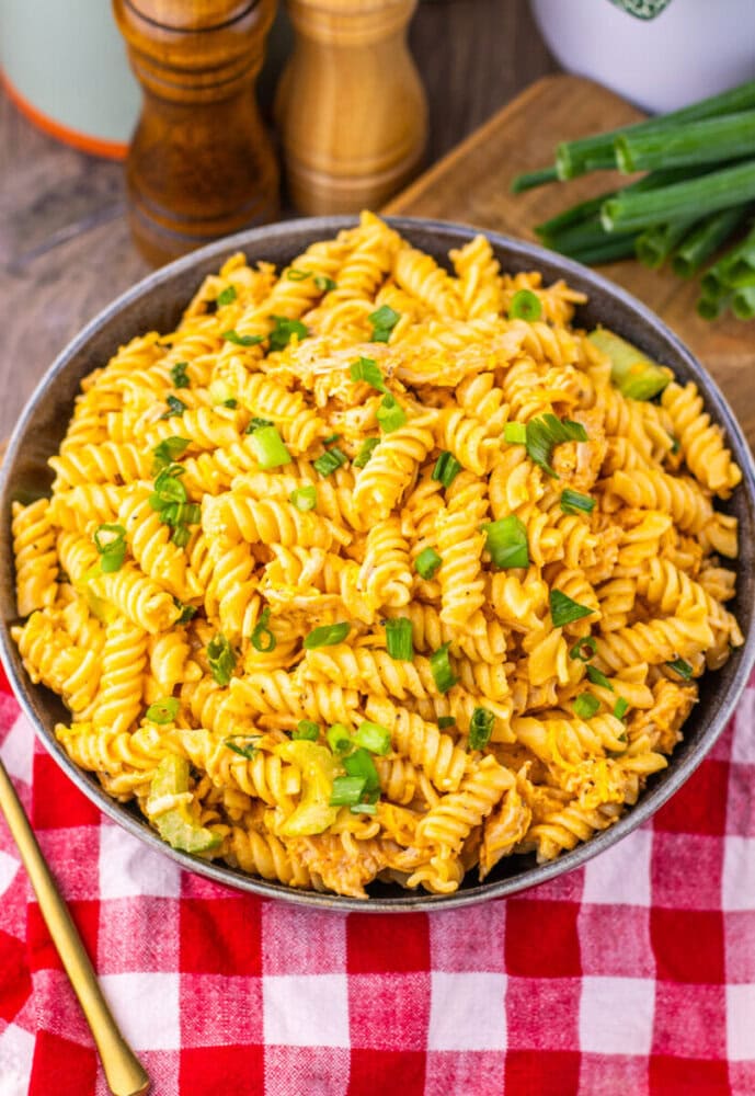 A bowl of creamy rotini pasta garnished with chopped green onions, on a red and white checkered cloth.