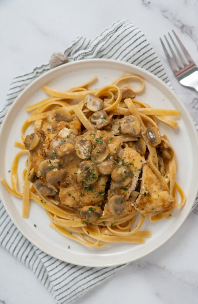 Plate of chicken and mushrooms in sauce served over fettuccine pasta, on a striped napkin with a fork nearby.