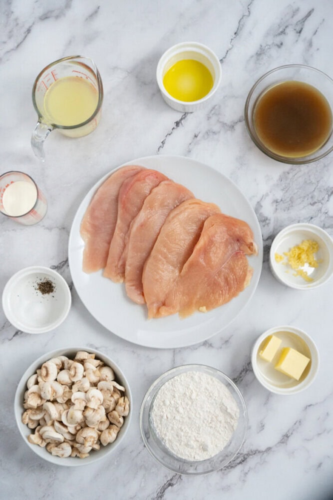 Raw chicken breasts on a plate, surrounded by small bowls of mushrooms, flour, butter, broth, cream, and seasonings.
