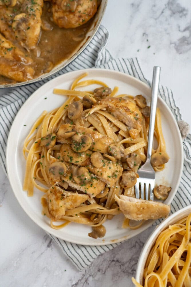 Plate of pasta topped with chicken and mushrooms in a creamy sauce, with a fork and striped napkin nearby.