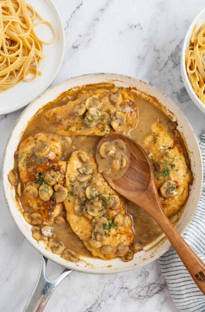 Chicken breasts in mushroom sauce in a skillet, with a wooden spoon; pasta in bowls on the side.