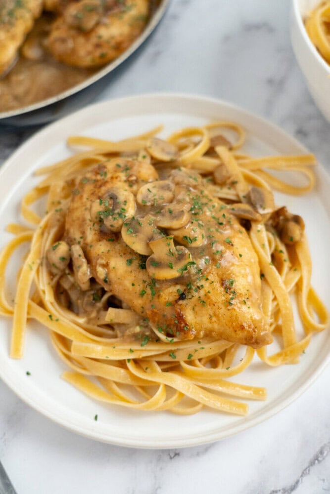 Chicken breast with mushroom sauce served over fettuccine pasta on a white plate, garnished with herbs.