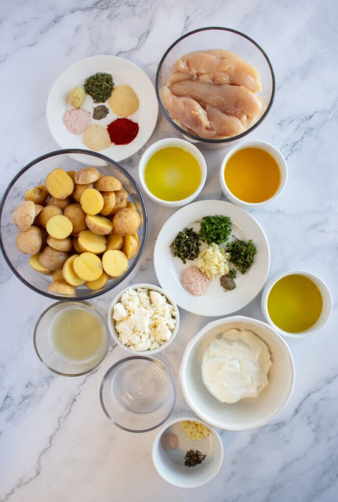 Bowls of ingredients including raw chicken, sliced potatoes, herbs, spices, feta, oil, yogurt, and broth on a marble surface.