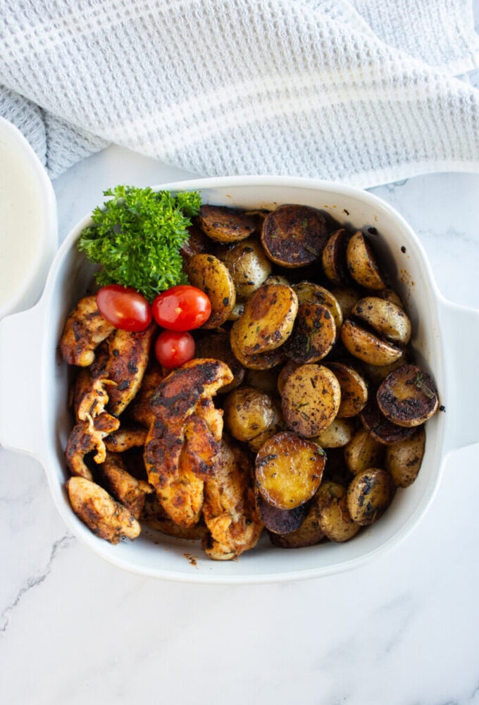 A white dish with roasted potato slices, grilled chicken, cherry tomatoes, and parsley garnish.