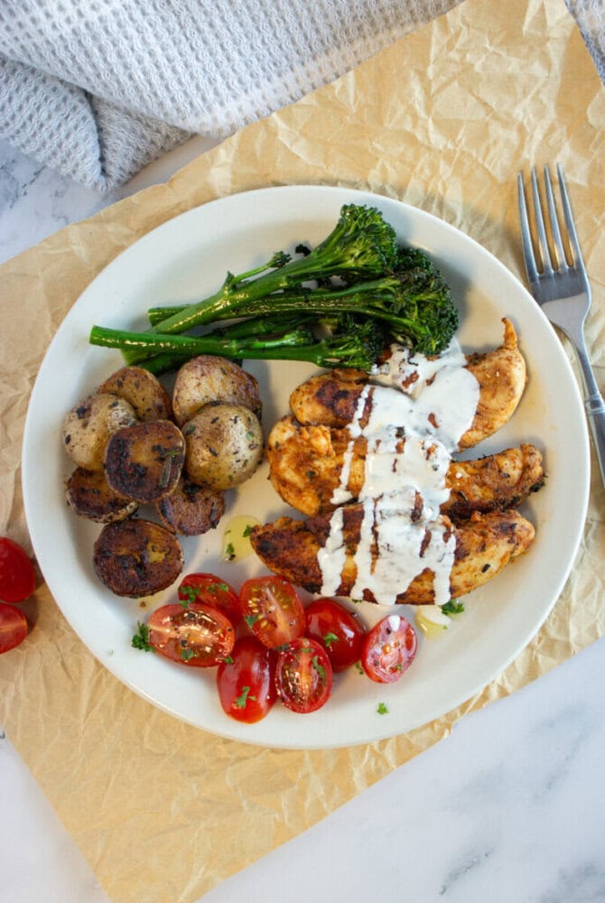 A plate with grilled chicken, roasted potatoes, broccolini, and cherry tomatoes, topped with white sauce.
