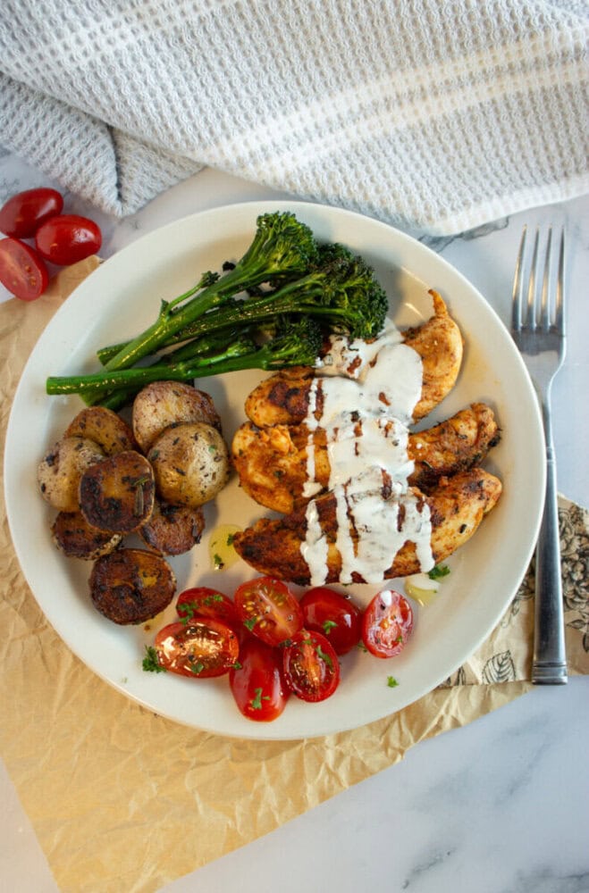 A plate with grilled chicken, roasted potatoes, broccolini, and cherry tomatoes, topped with a white sauce.
