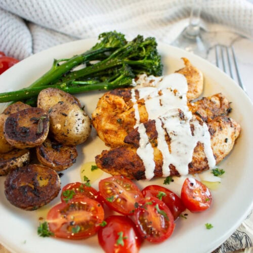 A plate with grilled chicken drizzled with sauce, roasted potatoes, broccolini, and sliced cherry tomatoes.