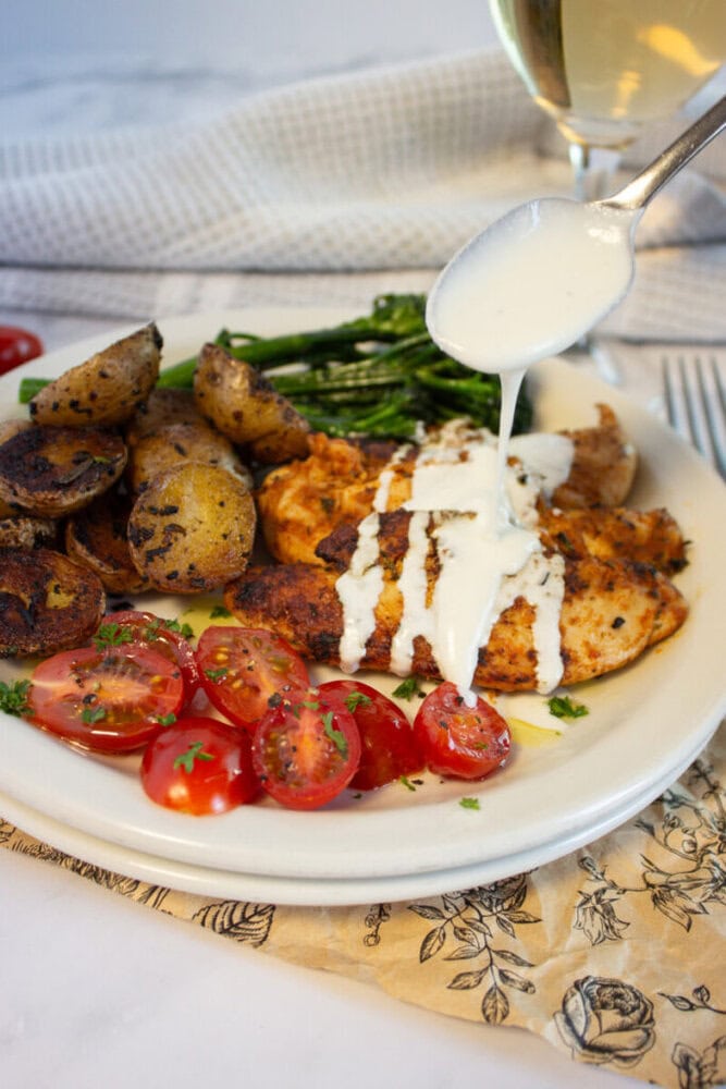 A spoon drizzles white sauce over grilled chicken with potatoes, green beans, and tomatoes on a plate.